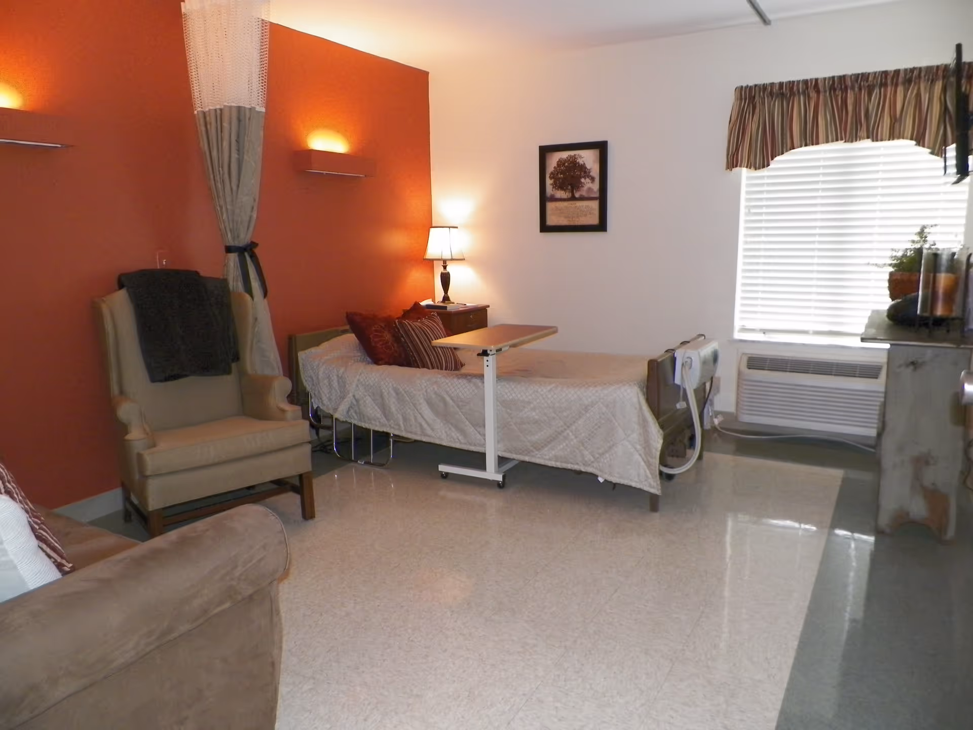 A senior living facility room with a single bed covered in a beige quilt, a small overbed table, a beige armchair with a dark throw draped over it, a brown sofa, a nightstand with a lamp, a framed picture on the wall, a window with blinds and a striped valance, and a heating/cooling unit below the window. The walls are painted white and burnt orange, and the floor is a shiny light-colored tile.