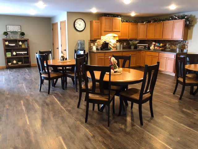A spacious dining area with round wooden tables and black chairs arranged on a wood floor. In the background, there is a kitchen area with wooden cabinets, a countertop, and a water dispenser. The room is well-lit with ceiling lights, and there are decorative plants and a clock on the wall.