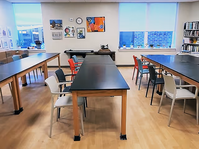 Communal activity room with long black-topped tables, mixed-color chairs, artwork on the walls, and large windows.
