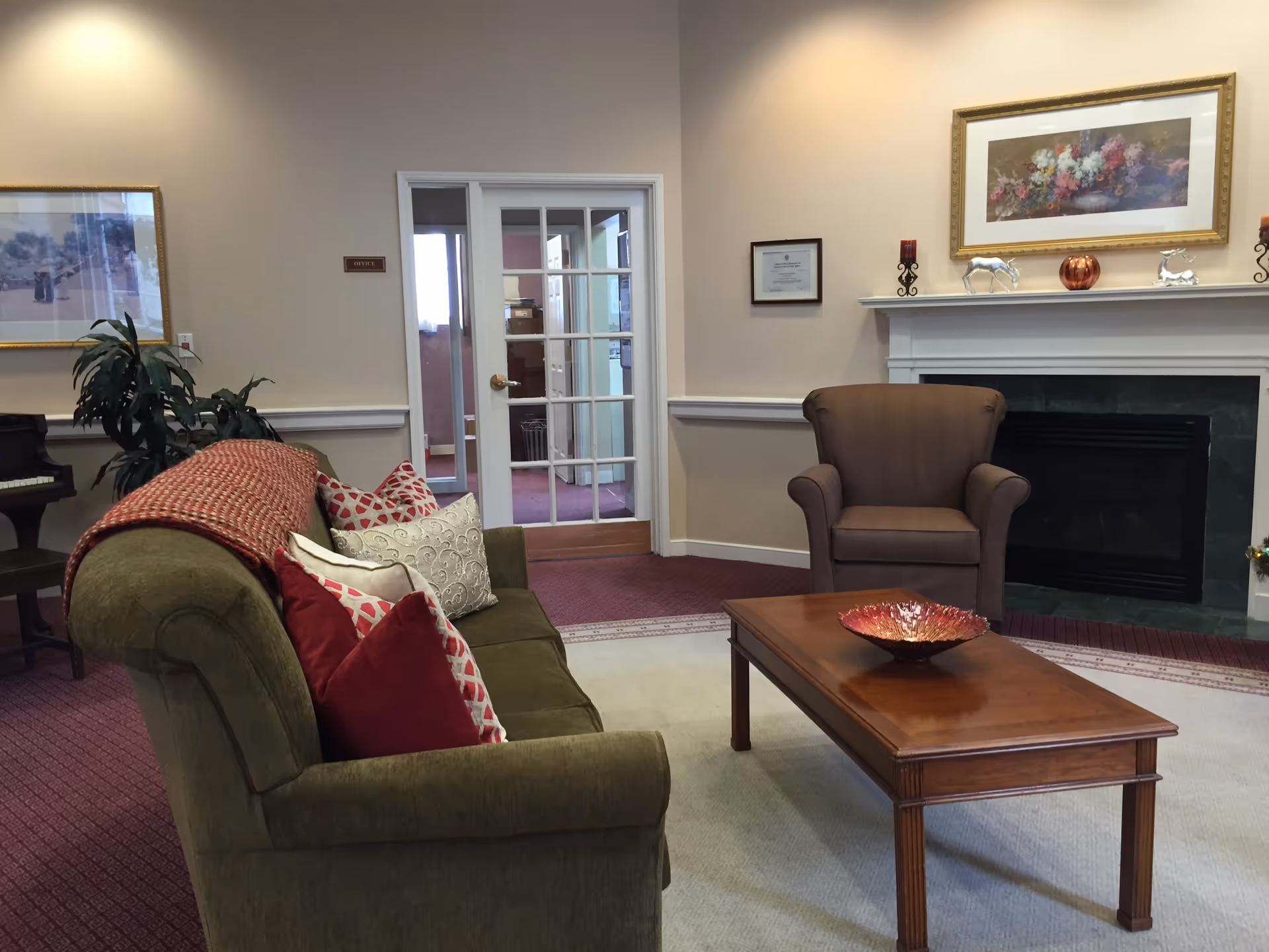 A cozy living room area with an olive green sofa adorned with red and white patterned pillows and a red throw blanket. In front of the sofa is a wooden coffee table with a decorative bowl. To the right is a brown armchair positioned near a white fireplace mantel decorated with candles, small silver animal figurines, and a framed floral painting above. There is a glass-paneled door labeled 'Office' in the background, and a piano partially visible on the left side of the image.