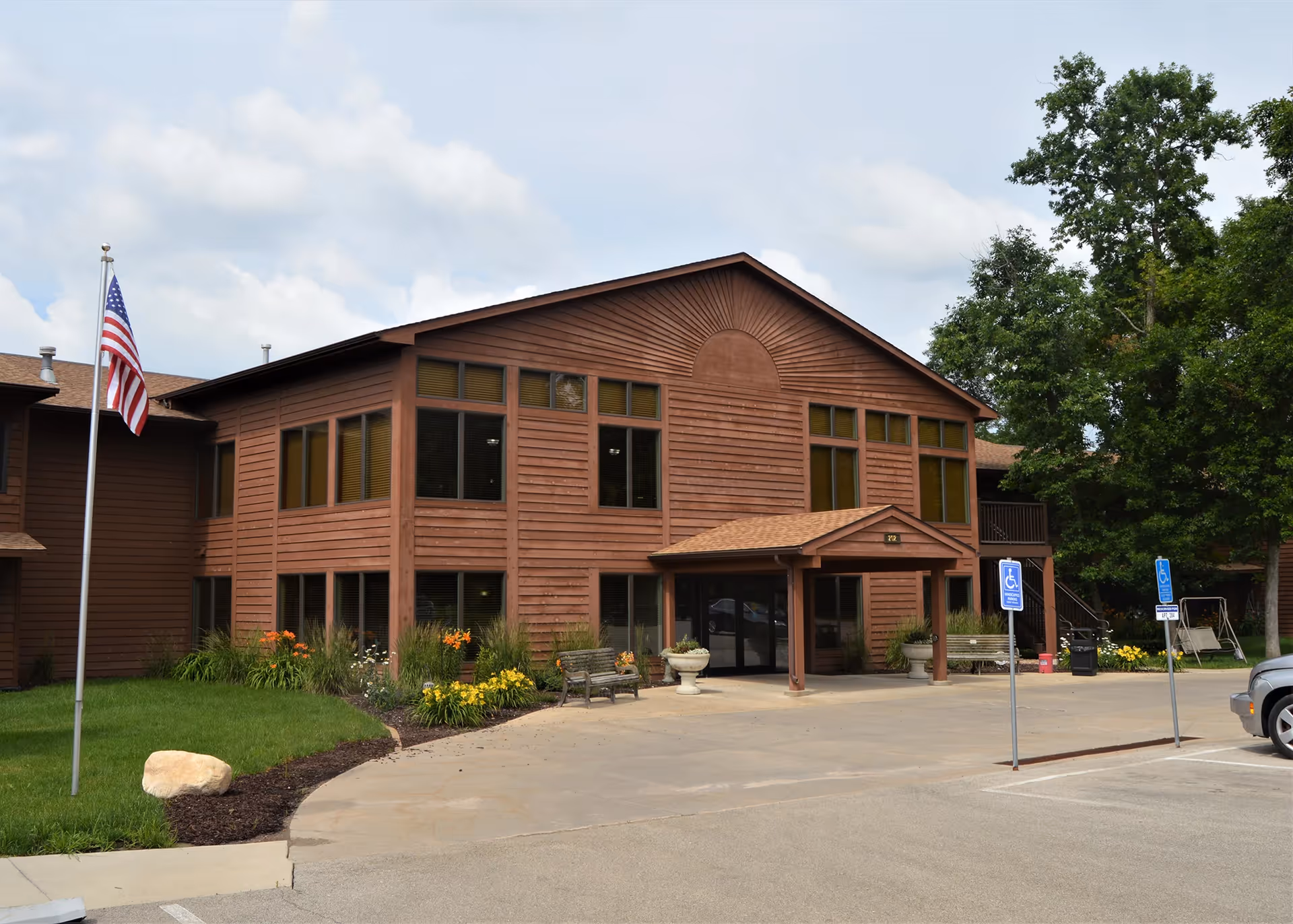 Front exterior of a two-story brown wood-clad senior living building with a covered entrance, American flag, landscaping and handicapped parking spaces.