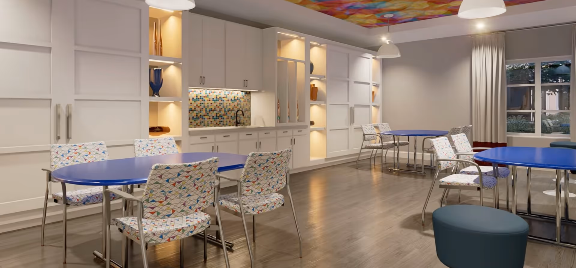 A bright and modern common area with blue tables and colorful patterned chairs. The room features white cabinetry with built-in shelves displaying decorative vases, a mosaic tile backsplash above a sink, wood flooring, large windows with curtains, and pendant lighting hanging from a ceiling with a colorful geometric design.