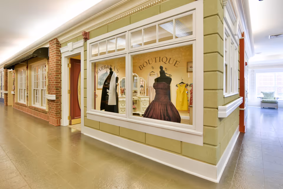 Interior hallway of a senior living facility featuring a boutique with mannequins dressed in clothing displayed behind large windows. The boutique is labeled 'Barbara's Boutique'. The hallway has brick and painted walls with multiple doors and windows, leading to a bright seating area in the background.