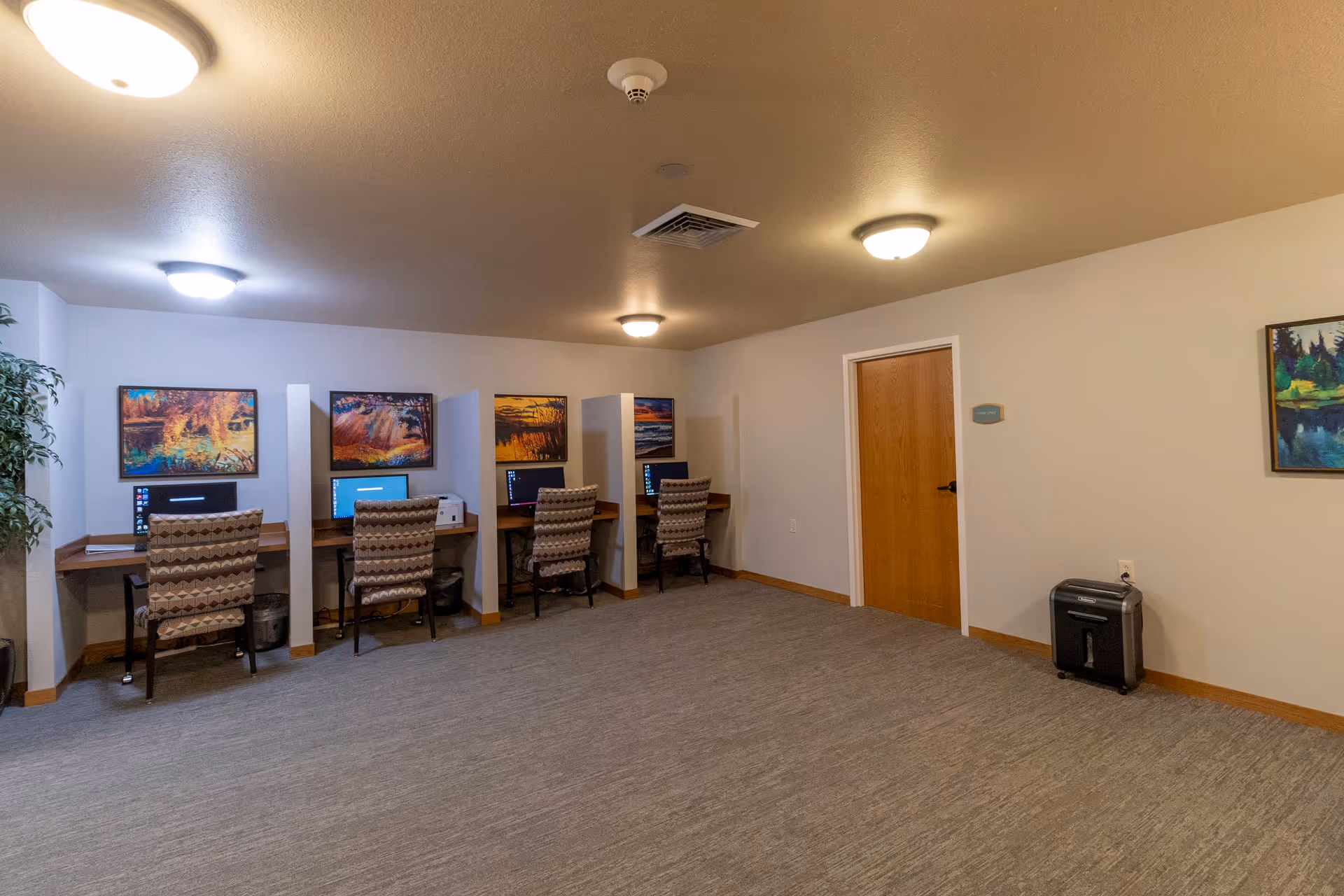 A quiet computer room with four individual workstations, each equipped with a desktop computer and a patterned chair. The walls are decorated with colorful landscape paintings, and the room has a beige carpet and ceiling lights. There is a wooden door and a paper shredder against the far wall.