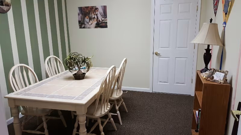 A dining room with a rectangular table featuring a tiled surface and six wooden chairs. The walls are painted light green with one wall having vertical green and white stripes. There is a small plant centerpiece on the table. On the right side, there is a wooden bookshelf with a lamp and some decorative items on top. A closed white door is visible at the back of the room.
