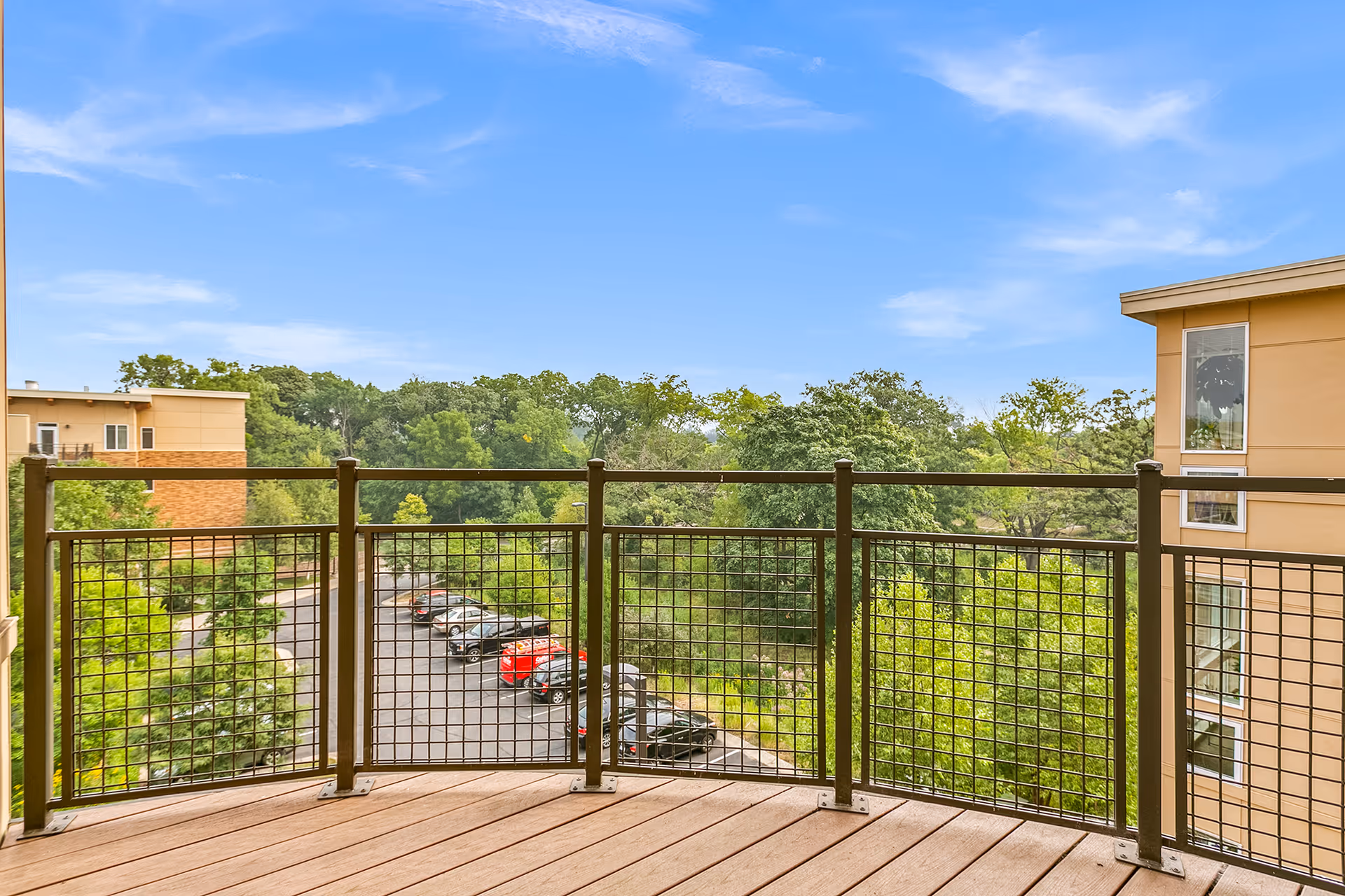 View from a balcony with wooden flooring and metal railing overlooking a parking lot with several cars and green trees under a blue sky with some clouds.