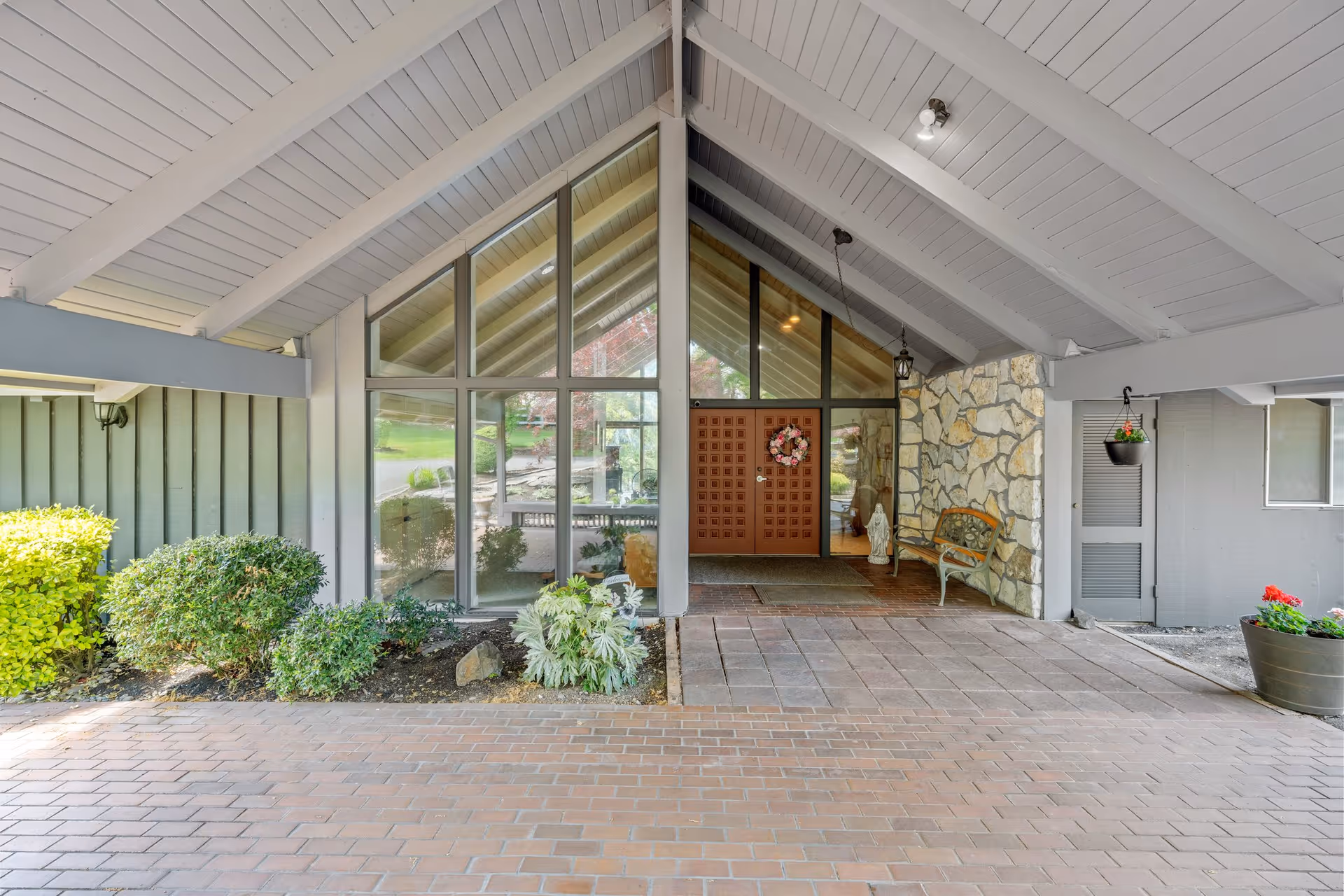 Covered entrance area of a building with a high peaked ceiling, large glass windows, a wooden door decorated with a wreath, stone wall accents, a bench, and potted plants on either side.