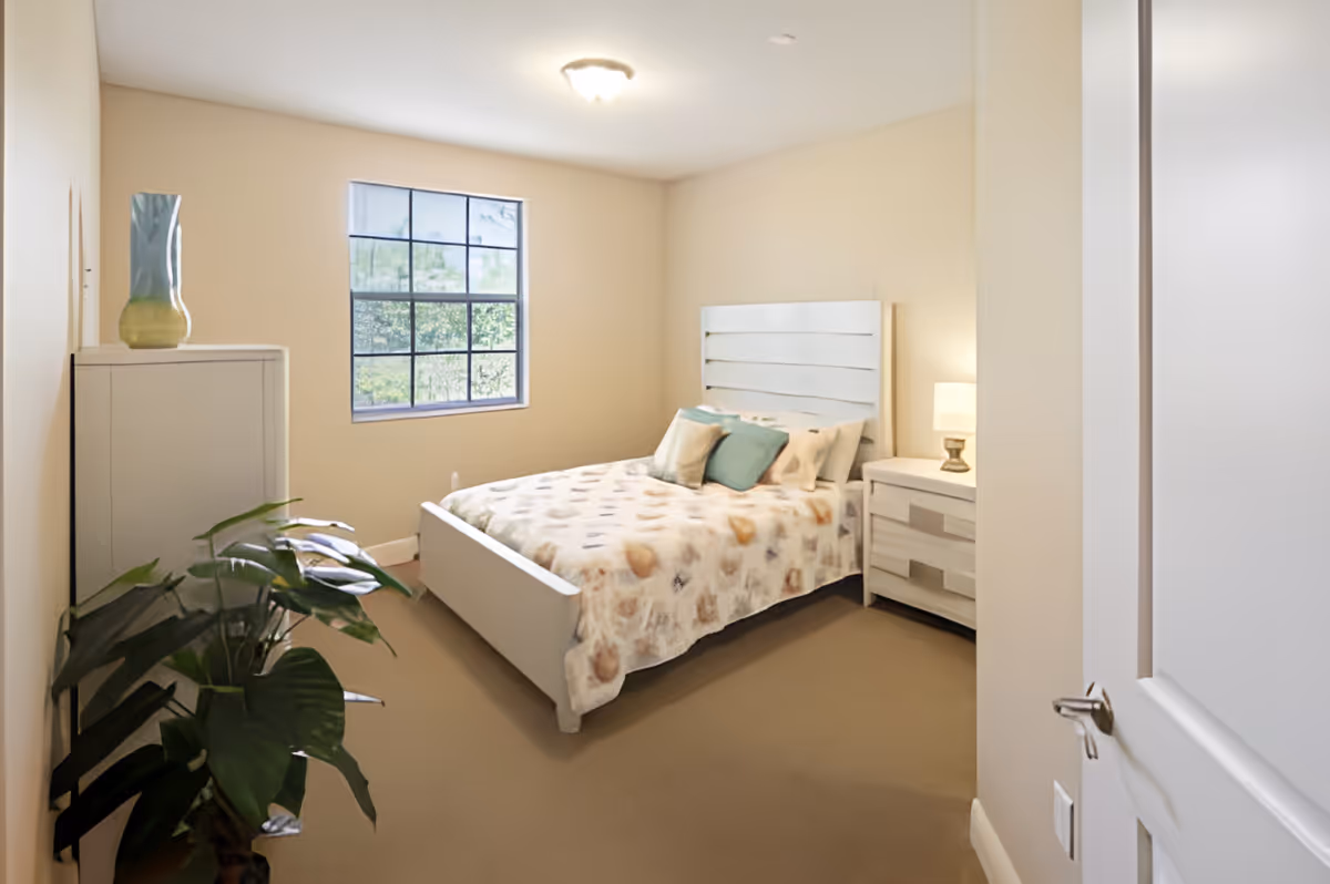 A cozy bedroom with a white bed frame and a bed covered with a patterned blanket and several pillows. There is a white nightstand with a lamp on the right side of the bed and a white dresser with a decorative vase on the left side of the room. A large window lets in natural light, and a green leafy plant is placed near the dresser. The walls are painted beige, and the carpet is light brown.