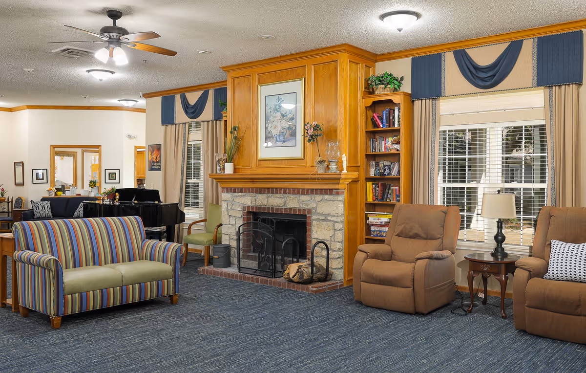 A cozy living room area in an assisted living facility featuring a stone fireplace with a wooden mantel, a striped loveseat, two brown recliners, a side table with a lamp, a bookshelf filled with books, and large windows with beige curtains and blue valances. The room has blue carpet flooring and ceiling fans with lights.