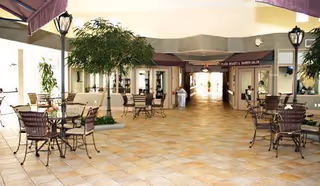 Indoor common area with tiled floor, several metal tables and chairs arranged around small trees and lamp posts, with a hallway leading to other rooms in the background.