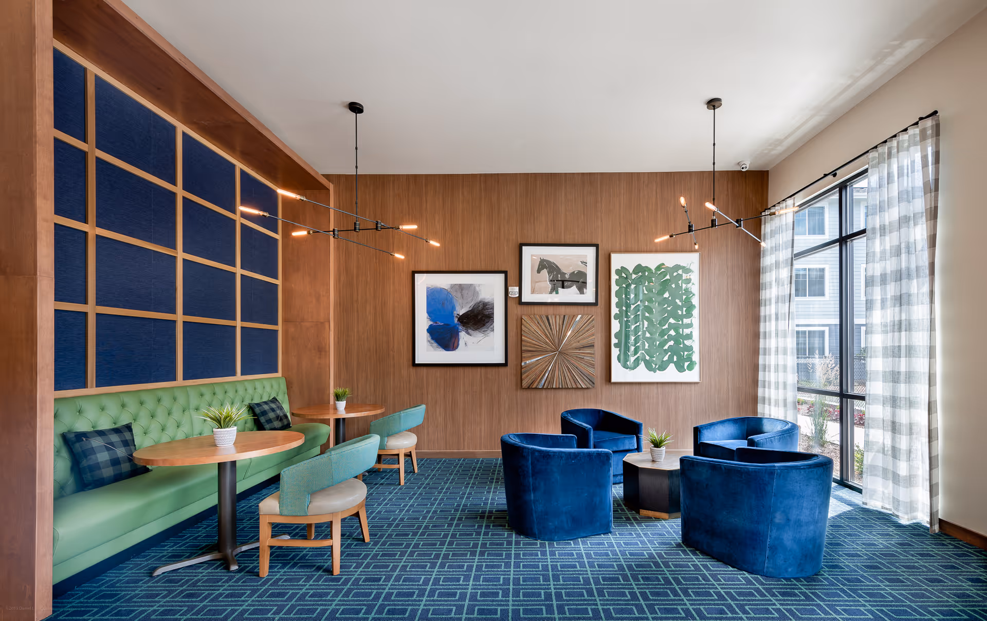 A modern lounge area with a green tufted bench along the left wall, two round wooden tables with green chairs, and four blue velvet armchairs arranged around a small black coffee table. The room features a blue patterned carpet, wooden panel walls with framed artwork, large windows with sheer checkered curtains, and contemporary pendant lights hanging from the ceiling.