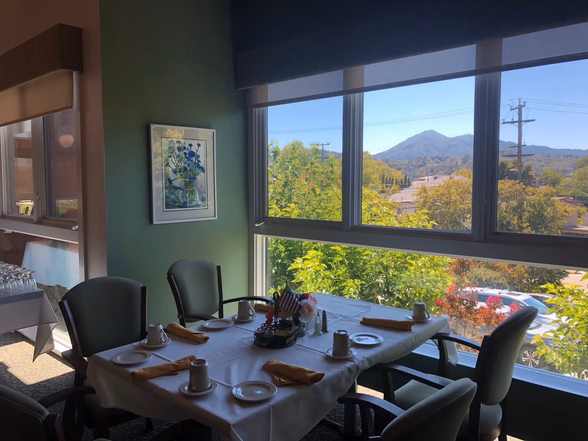 A dining table set for four with white tablecloth, yellow napkins, cups, and plates near large windows showing a view of trees, rooftops, and distant mountains under a clear blue sky. The room has green walls with a framed floral painting and additional tables with glassware in the background.
