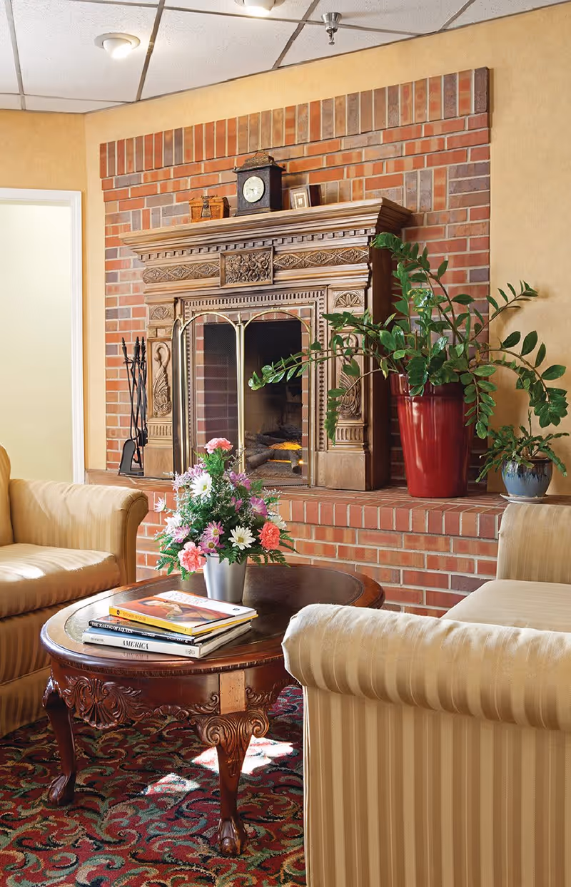 A cozy living room area featuring a brick fireplace with an ornate wooden mantle. On the mantle are a clock and small decorative items. In front of the fireplace is a wooden coffee table with a floral arrangement and several books. Two beige striped upholstered armchairs are positioned around the table, and there are two potted plants on the fireplace hearth. The room has a patterned carpet and warm yellow walls.