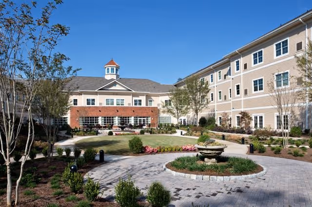 Landscaped courtyard with a central fountain and walkways in front of a multi-story beige senior living building under a clear blue sky.