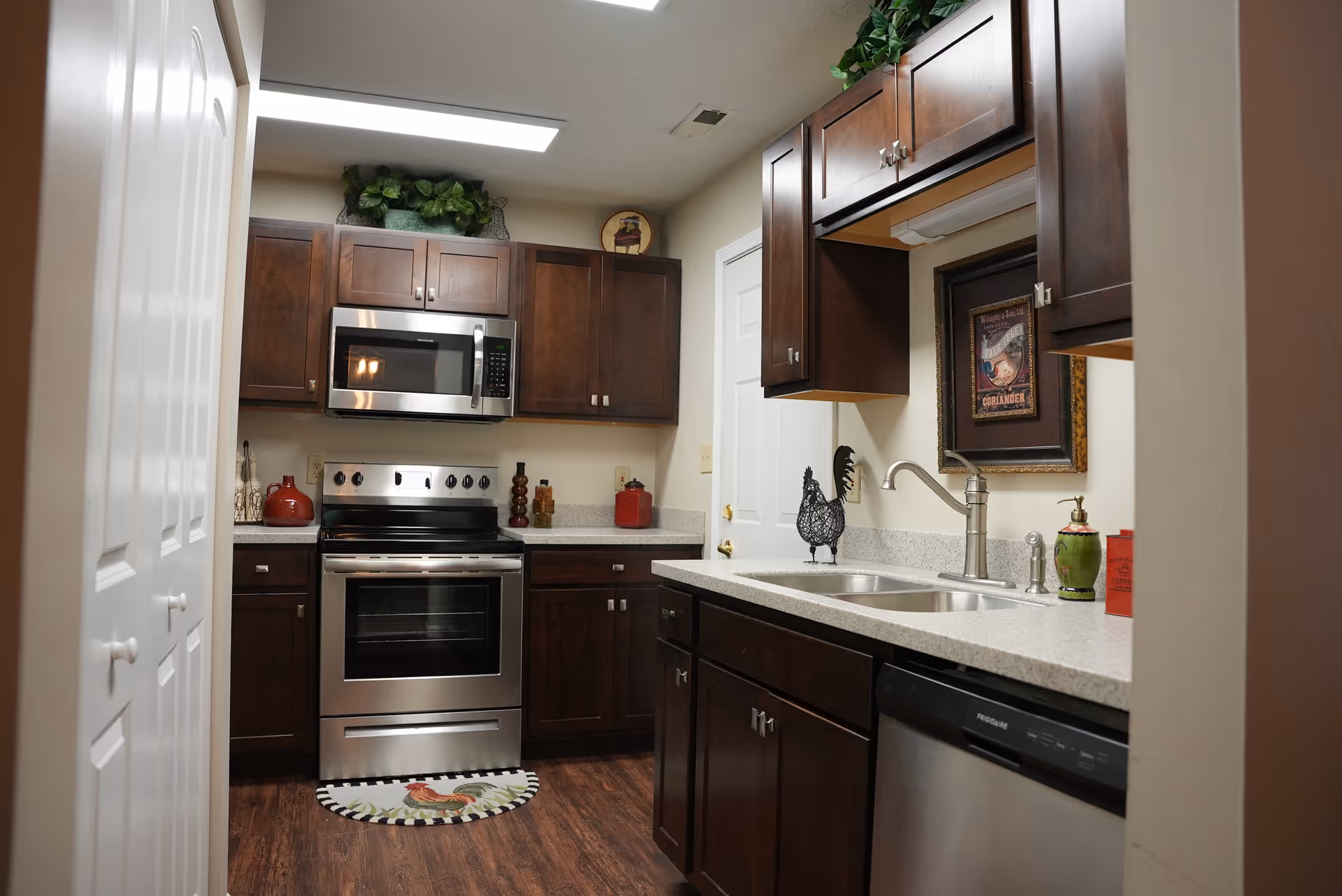 A modern kitchen with dark wooden cabinets, stainless steel appliances including a microwave, oven, and dishwasher. The kitchen has a double sink with a silver faucet, a decorative rooster figurine, and various kitchen items on the countertops. The floor is wooden, and there is a small round rug with a rooster design in front of the oven.