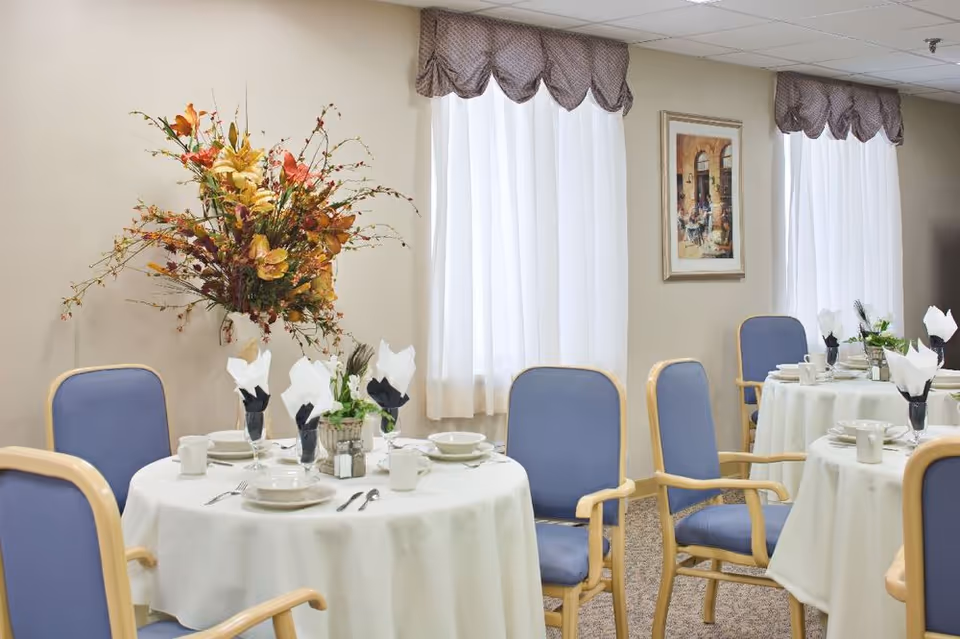 A dining room with round tables covered in white tablecloths, each set with white dishes, cups, silverware, and napkins folded in glasses. Blue cushioned wooden chairs surround the tables. There are two windows with white curtains and brown valances, a large floral arrangement on one table, and a framed painting on the wall.