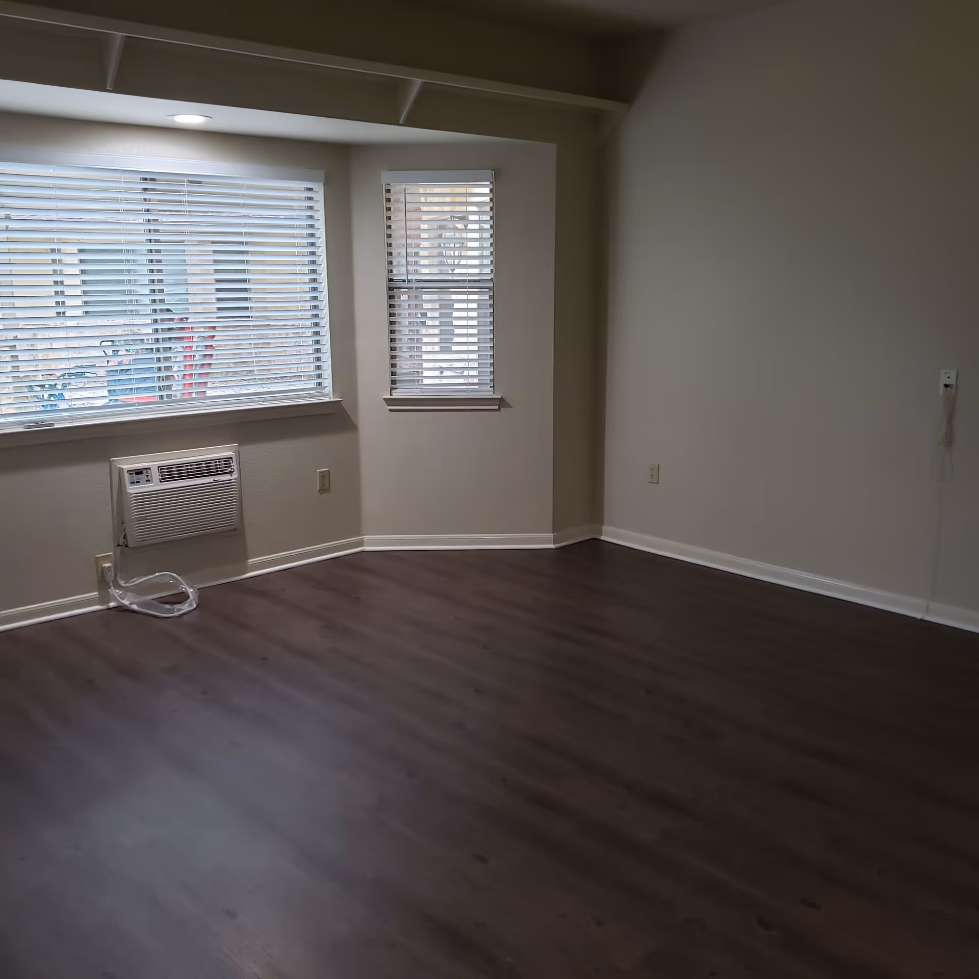Empty room with dark wood floors, beige walls, two windows with blinds and a wall air conditioner unit.