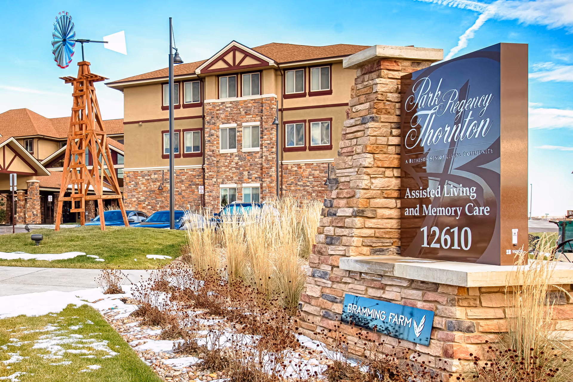 Exterior view of a senior living facility with a stone and brick building in the background, a wooden windmill structure on the lawn, and a large stone sign in the foreground that reads 'Park Regency Thornton Assisted Living and Memory Care 12610'. There is some snow on the grass and a clear blue sky with clouds.