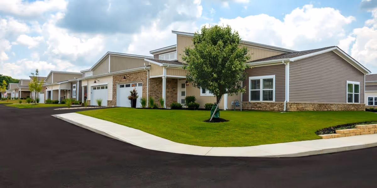 Exterior view of a modern single-story residential building with beige siding, stone accents, and attached garages. The building is surrounded by a well-maintained green lawn, a small tree, and a paved driveway under a partly cloudy sky.