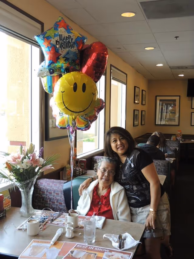 An elderly woman and a younger woman pose together at a restaurant booth next to birthday balloons and a vase of flowers.