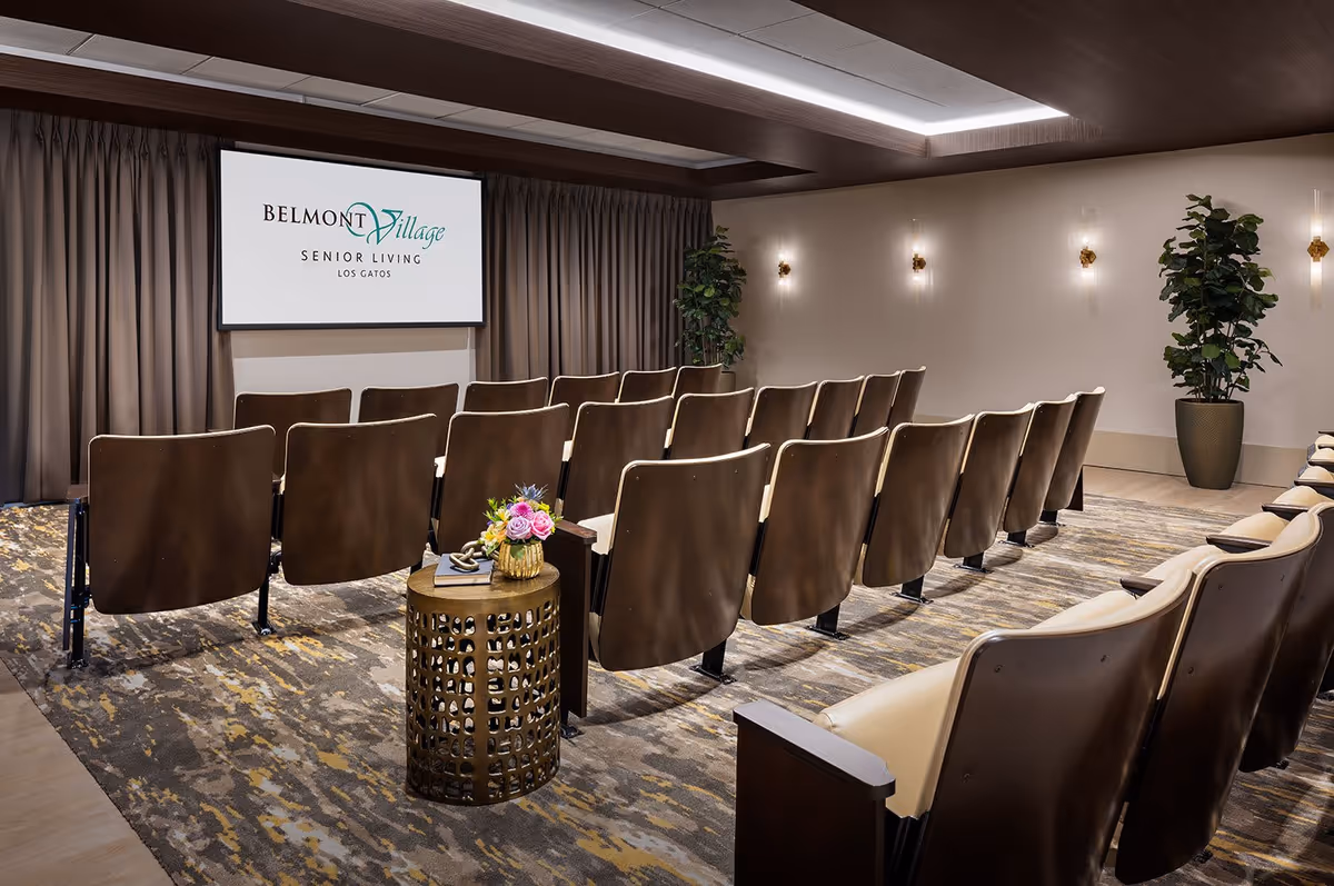 A small theater or presentation room with rows of brown cushioned chairs facing a large screen displaying the Belmont Village Senior Living Los Gatos logo. The room has a patterned carpet, beige walls with wall sconces, a potted plant in the corner, and a small round table with a flower arrangement and books.