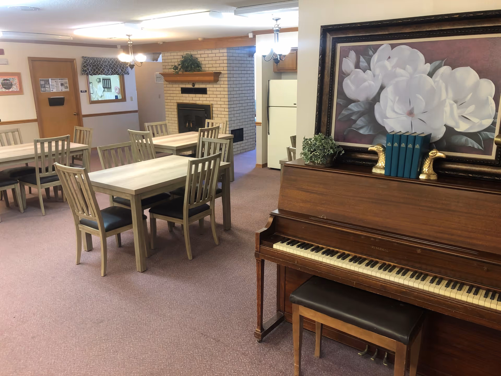 Interior view of a senior living facility common area with several wooden tables and chairs arranged on a carpeted floor. A wooden upright piano with a bench is in the foreground, topped with a framed painting of white flowers and decorative bookends holding blue books. In the background, there is a white brick fireplace with a wooden mantle and a white refrigerator partially visible behind a wall.
