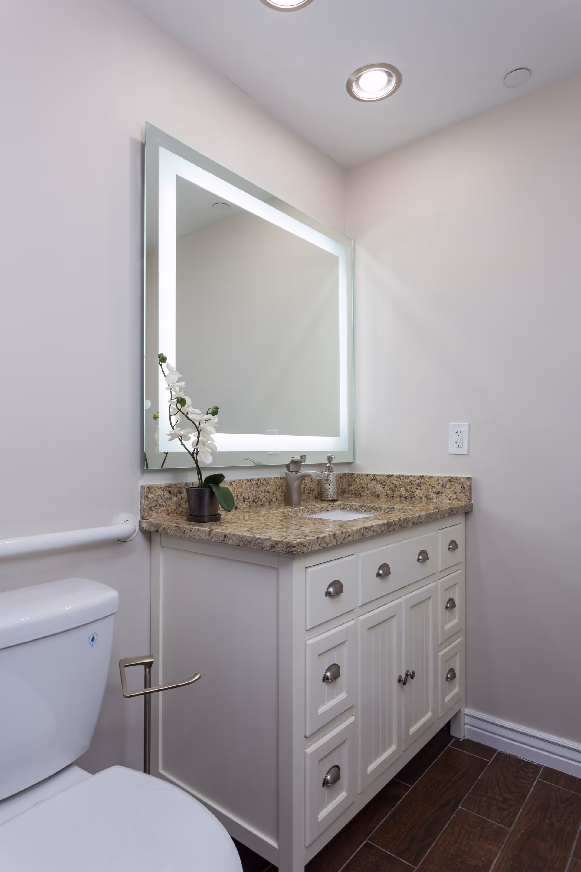 A clean and modern bathroom featuring a white toilet, a white vanity with granite countertop, a built-in sink, a soap dispenser, a small potted orchid plant, and a large illuminated mirror mounted on the wall. The floor has dark wood-like tiles and the walls are painted light beige.