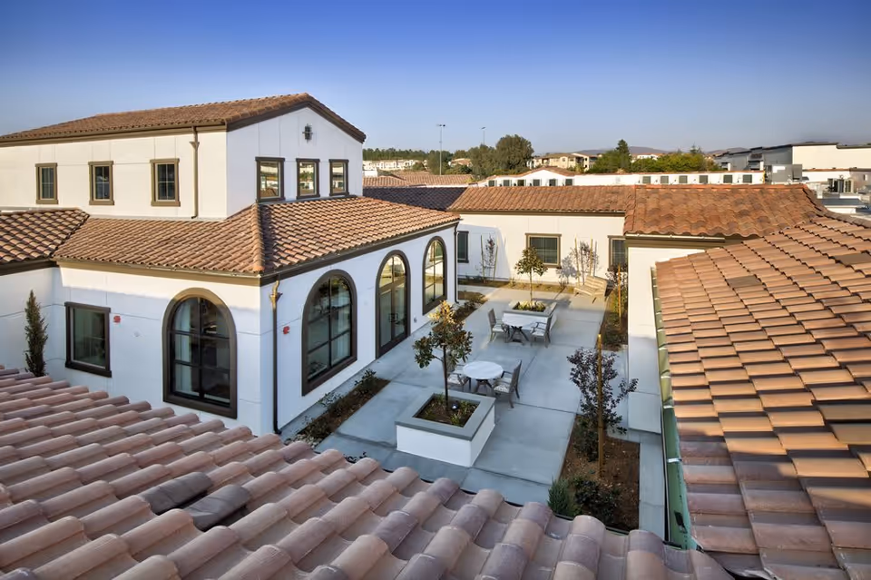 View of an outdoor courtyard area at Temecula Memory Care facility, featuring white buildings with terracotta tiled roofs, arched windows, small trees, and patio tables with chairs on a concrete pathway.