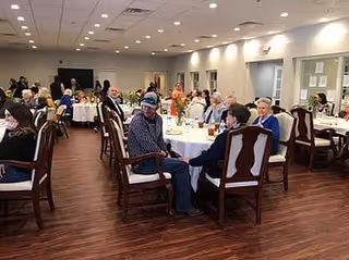 Communal dining/activity room with many residents seated at round tables on a wood floor under recessed ceiling lights.