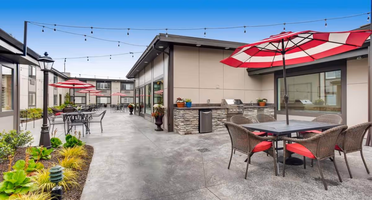 Outdoor patio area at Cascade Inn Assisted Living Community featuring multiple tables with red and white striped umbrellas, wicker chairs with red cushions, string lights overhead, a built-in stone grill, and surrounding greenery.