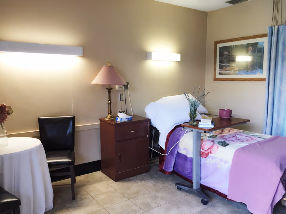 A cozy room with a single bed covered in floral and purple bedding, a bedside table with a lamp and tissue box, a small table with a vase of flowers, a black chair, and a wall-mounted light fixture. There is a framed picture on the wall and a curtain partially visible on the right side.