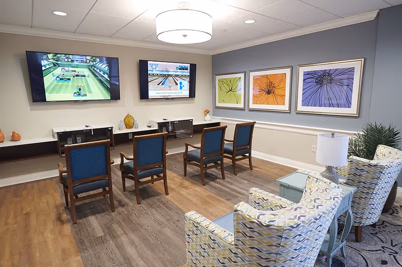 A cozy senior living common area with two wall-mounted TVs displaying video games, four wooden chairs with blue cushions facing the screens, two patterned armchairs with a small table and lamp between them, and three framed floral artworks on the wall.