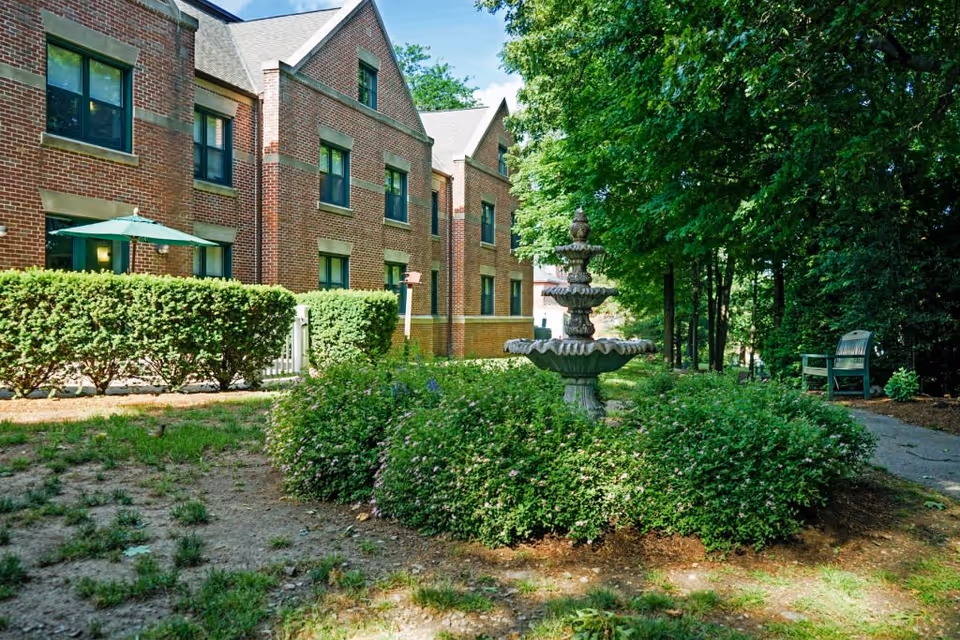 Outdoor garden area at Stone Rehabilitation And Senior Living featuring a multi-tiered stone fountain surrounded by green bushes, a brick building with multiple windows in the background, a green umbrella over a patio area, and a bench along a shaded pathway under tall trees.