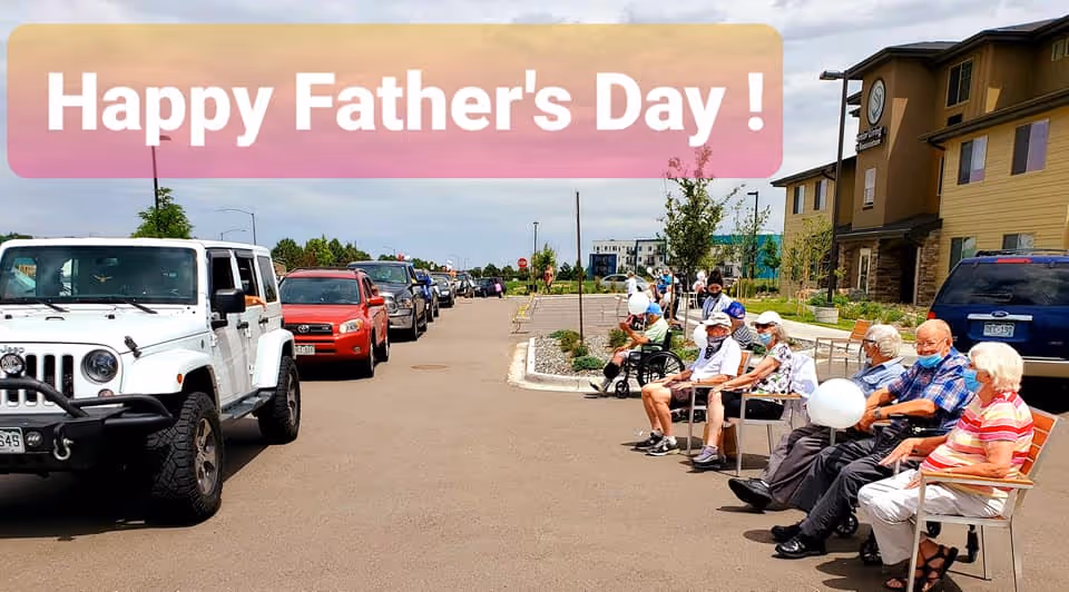 A group of elderly people sitting on benches outside a senior living facility, wearing masks and holding white balloons, with a line of cars passing by on the street. A banner at the top says 'Happy Father's Day!'