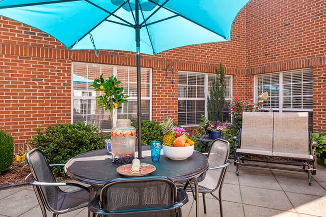 Outdoor patio area with a round black metal table and four chairs under a large blue umbrella. On the table, there is a bowl of fruit, a decorative pitcher, a blue glass, and a plate with a rolled napkin. The patio is surrounded by red brick walls with windows and various green plants and flowers.