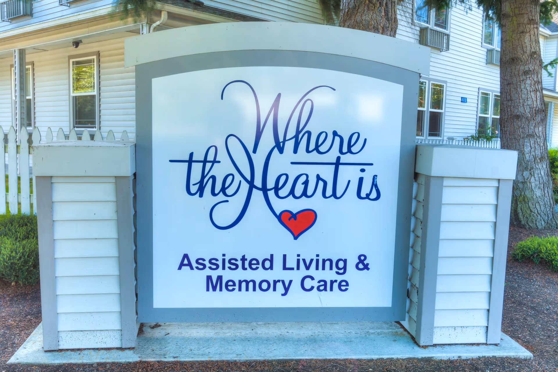 A large outdoor sign for 'Where the Heart is' Assisted Living & Memory Care, positioned in front of a white building with windows and surrounded by trees and shrubs.
