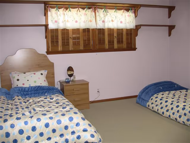 A simple bedroom with two single beds, each with blue and beige polka dot bedding and blue blankets. There is a wooden nightstand between the beds with a small mirror and an alarm clock on it. Above the nightstand is a window with wooden shutters and a floral curtain. The walls are light-colored and there are wooden shelves mounted on the walls.