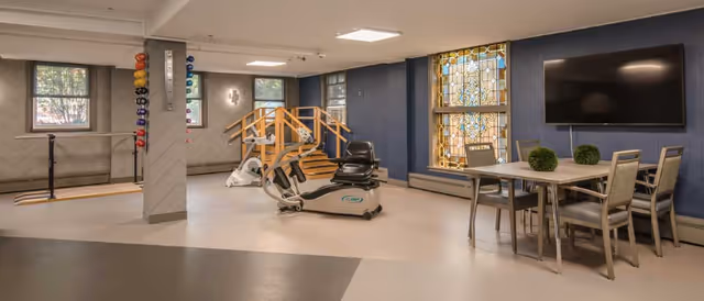 A spacious exercise room with light-colored flooring and blue walls. The room features various fitness equipment including a recumbent exercise bike, a stationary bike, and a set of colorful dumbbells mounted on a vertical rack attached to a pillar. There is a small wooden staircase with handrails leading to a raised platform. On the right side, there is a table with four chairs and a large flat-screen TV mounted on the wall above it. A large stained glass window adds decorative detail to the room.