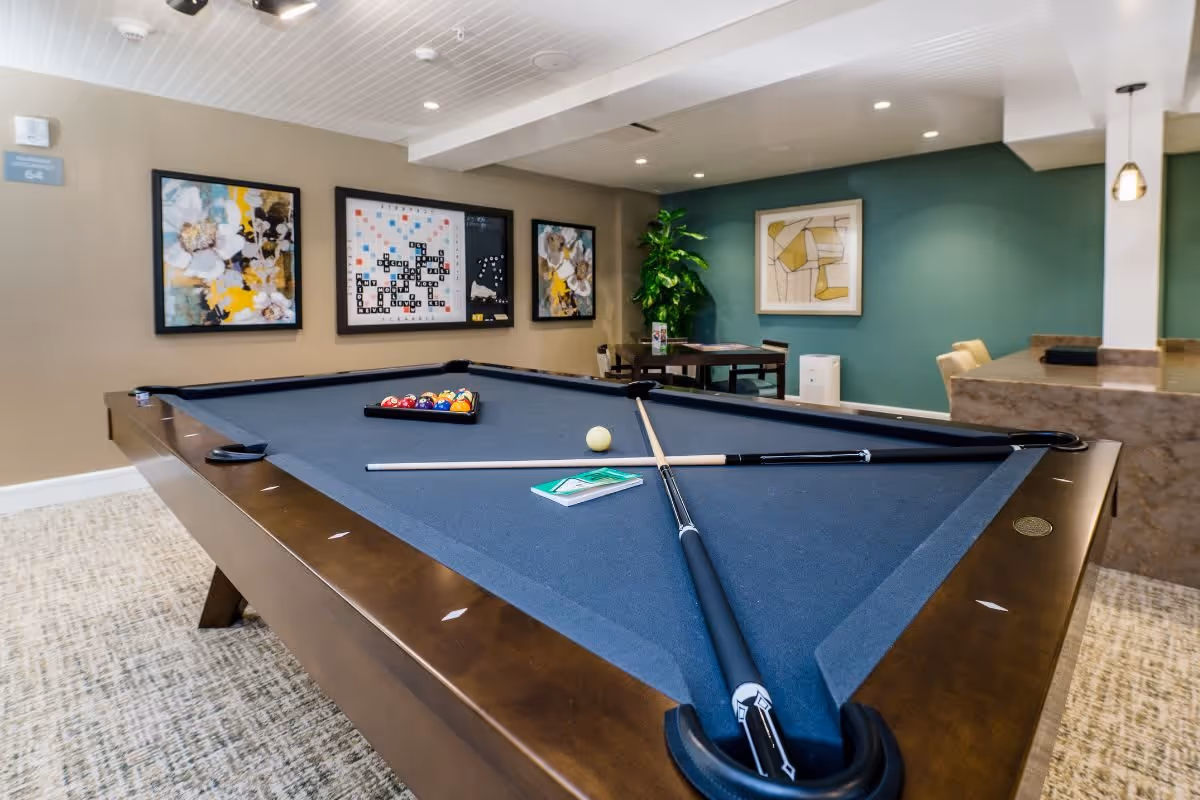 A recreational room featuring a pool table with a blue felt surface, pool cues, a cue ball, and a rack of billiard balls. The room has framed artwork on the walls, a small table with chairs, a green accent wall, and a potted plant. There is also a countertop with bar stools on the right side.
