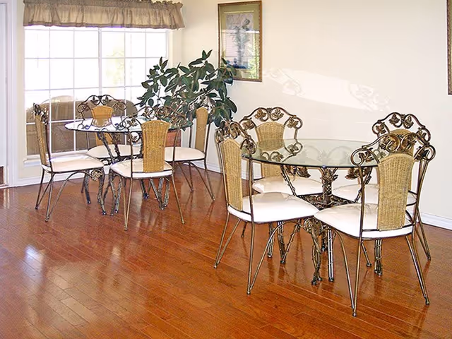 A dining area with two glass-top tables, each surrounded by four ornate metal chairs with cushioned seats and wicker backs. The room has wooden flooring, a large window with a valance, and a green potted plant in the corner.