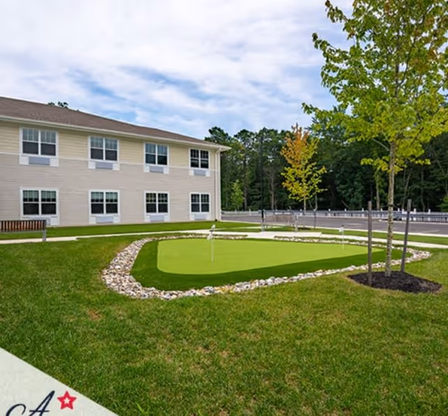 Outdoor putting green area with small flags surrounded by rocks and grass, adjacent to a two-story building with multiple windows. Trees and a parking lot are visible in the background under a partly cloudy sky.
