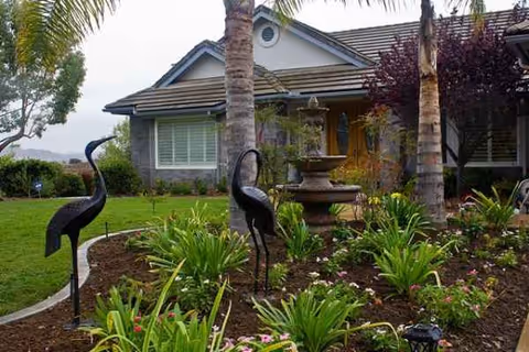 Front yard with tropical landscaping, a fountain, and a single-story senior living facility facade.