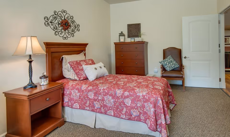 A bedroom with a single bed covered in a red floral bedspread, a wooden headboard, and multiple pillows. Next to the bed is a wooden nightstand with a lamp and a small decorative item. Across from the bed is a wooden chest of drawers with decorative items on top, and a brown leather armchair with a patterned cushion. The room has beige walls and carpeted flooring, with a white door partially open showing another room.