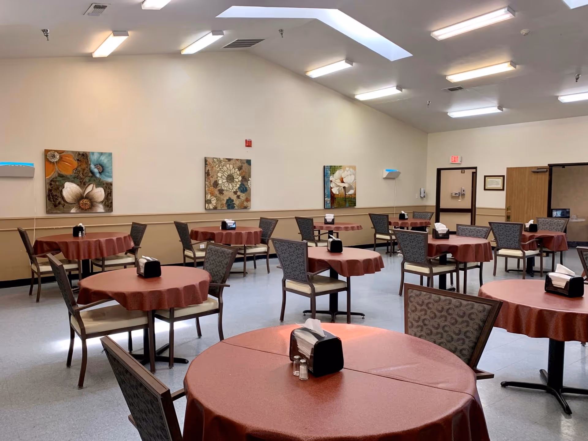 A dining room with multiple round tables covered with maroon tablecloths, each table surrounded by chairs with patterned upholstery. The room has beige walls adorned with floral paintings and a ceiling with fluorescent lights and skylights. Napkin holders and salt and pepper shakers are placed on each table.