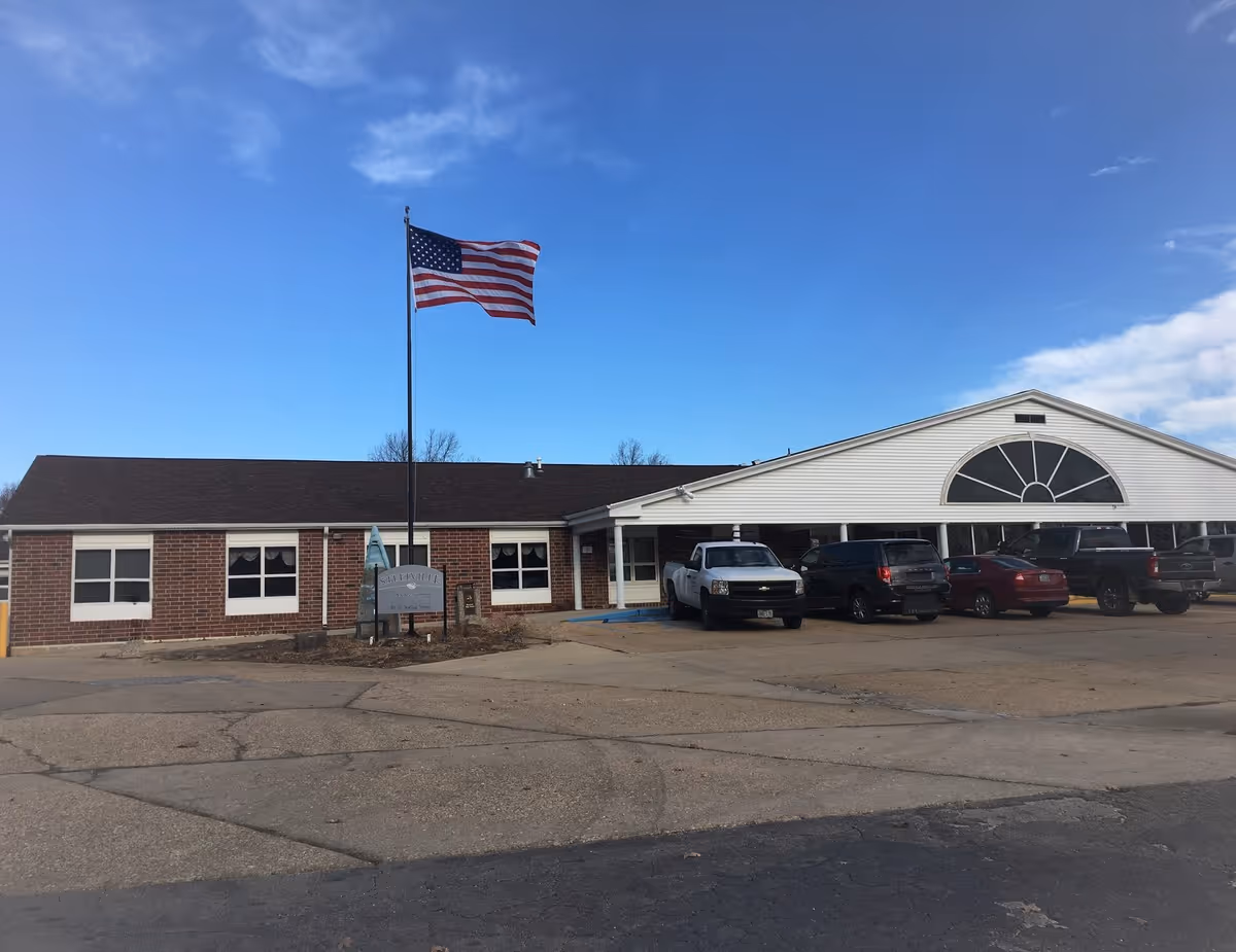 Exterior view of Steelville Senior Living facility showing a single-story brick and white building with several parked vehicles in front. An American flag is flying on a flagpole near the entrance under a clear blue sky.