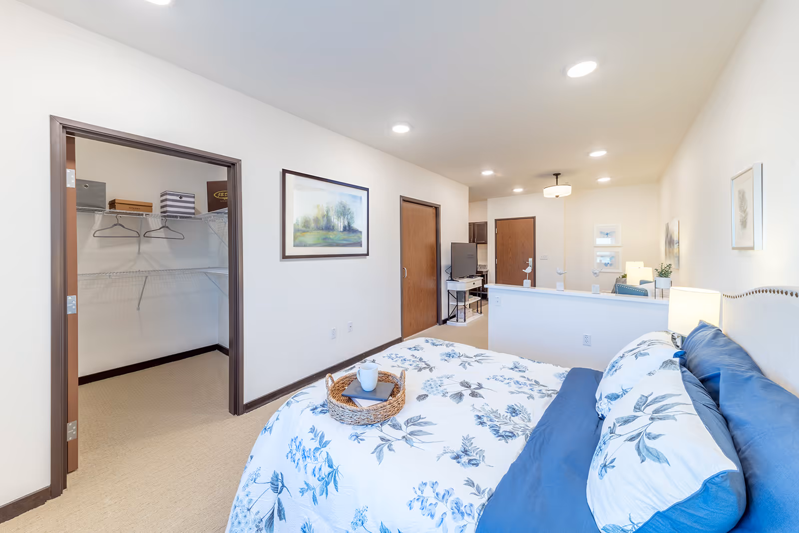 Bright furnished bedroom with floral bedding, a tray on the bed, an open walk-in closet, and a view into the living area.