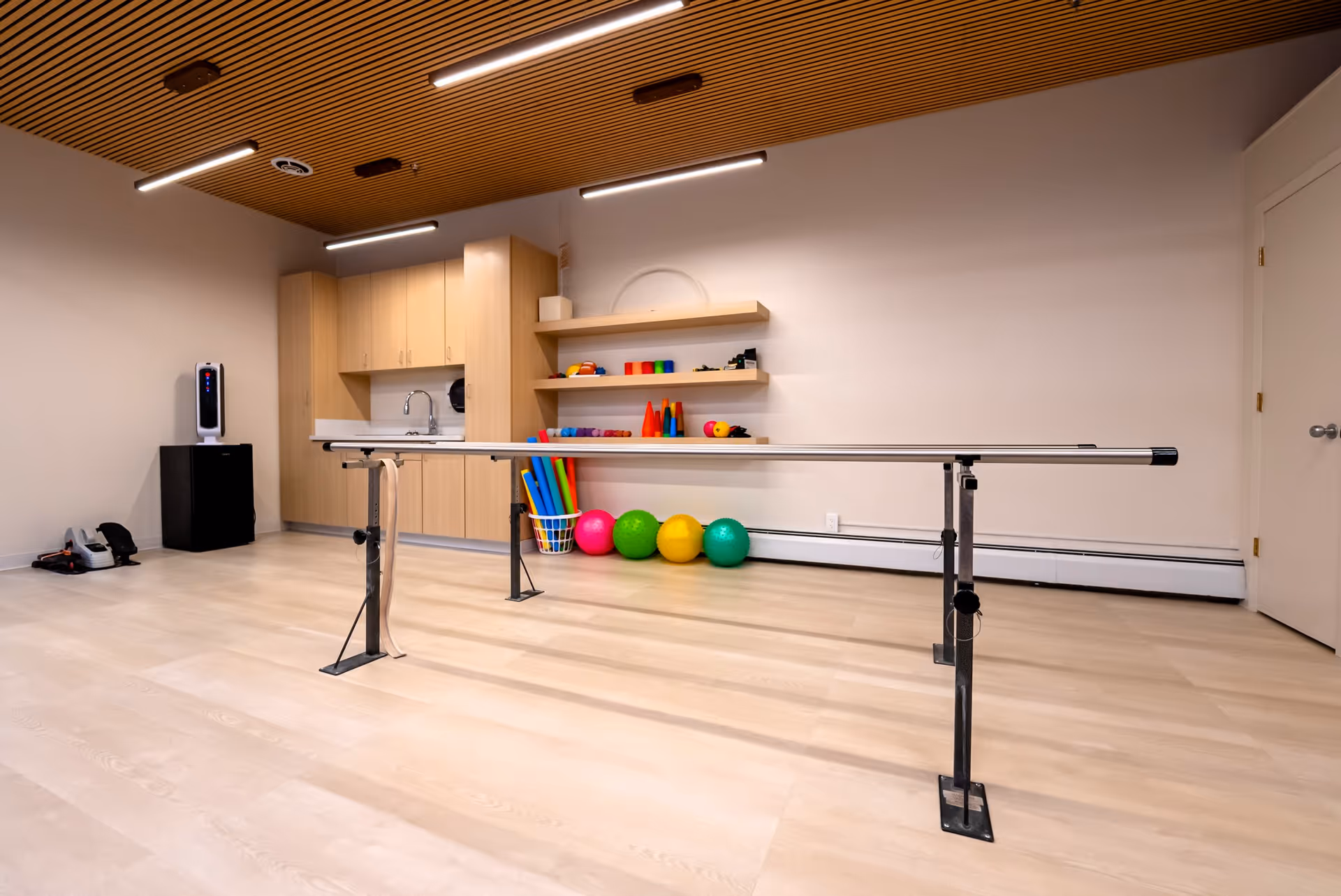 Rehabilitation/therapy room with parallel bars, colorful exercise balls, cabinets, shelves and a sink.