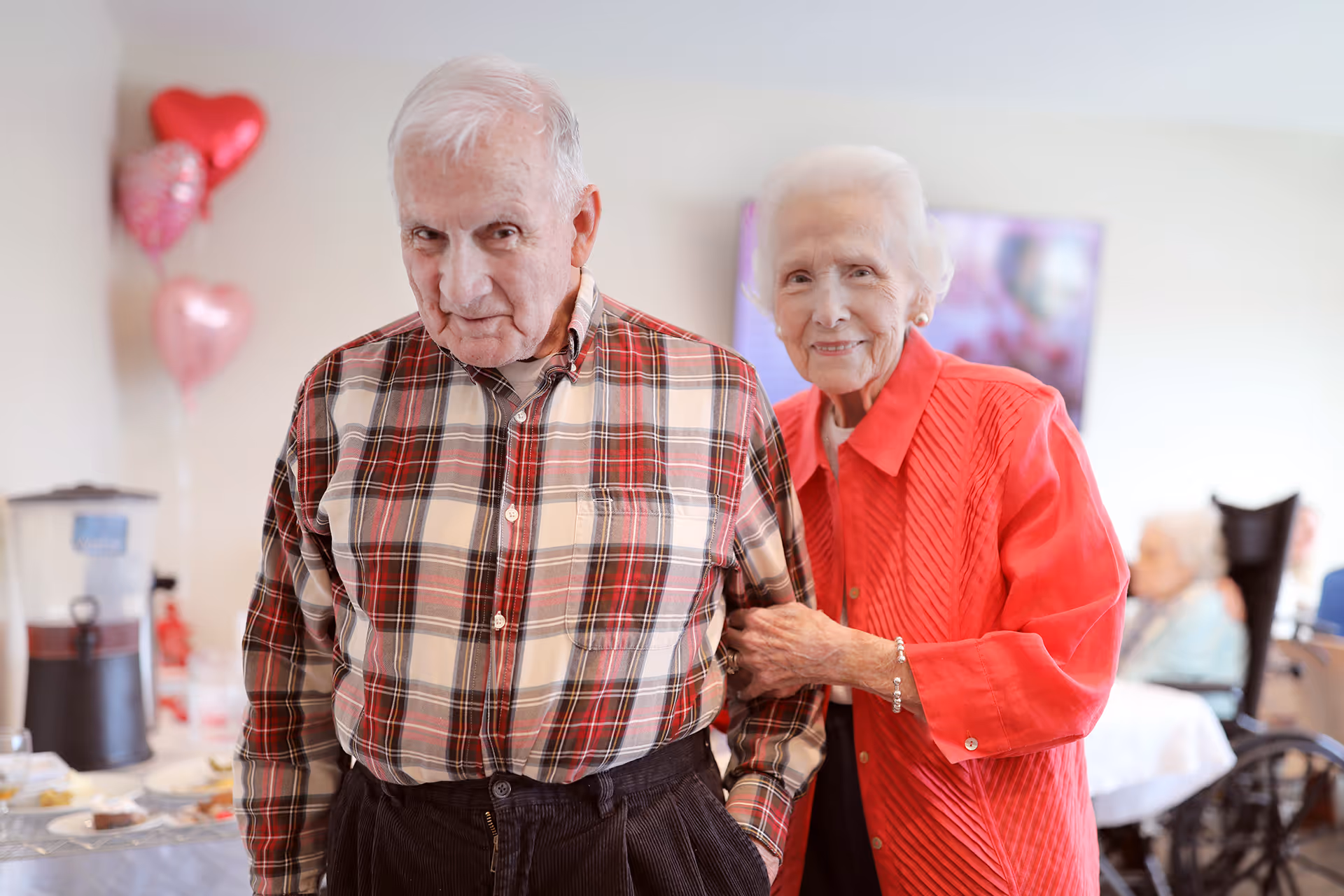 An elderly man and woman smiling and posing together in a decorated common room with balloons and tables in the background.