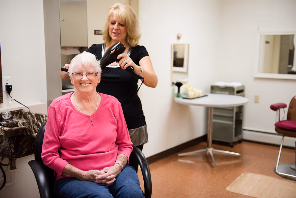 An elderly woman with white hair and glasses is sitting in a salon chair smiling while a hairstylist blow-dries her hair in a well-lit room with a sink, mirrors, and salon furniture.