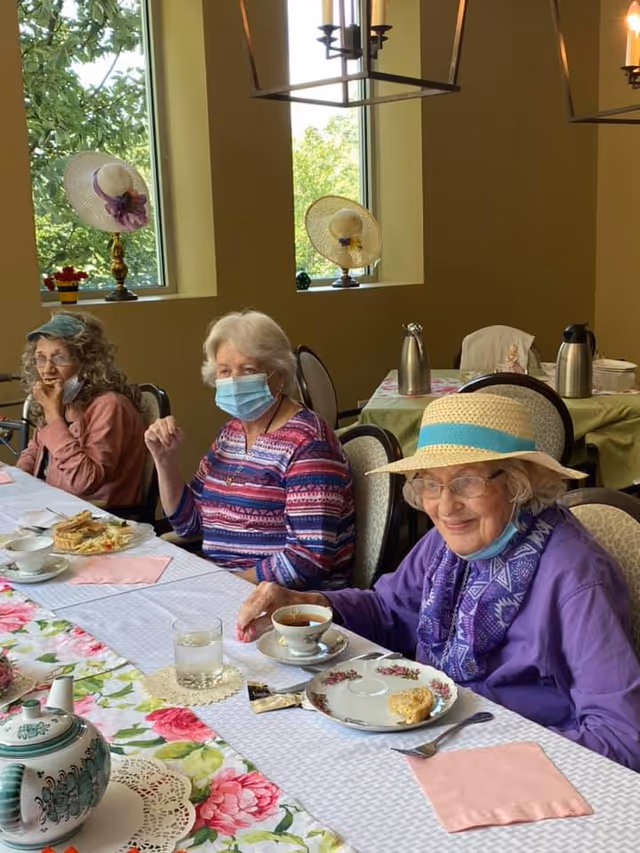 Three elderly women sitting at a dining table in a well-lit room with large windows. Two women are wearing hats and one woman is wearing a face mask. The table is set with floral tablecloths, teacups, plates with food, and a teapot. Decorative hats are displayed on the window sills behind them.